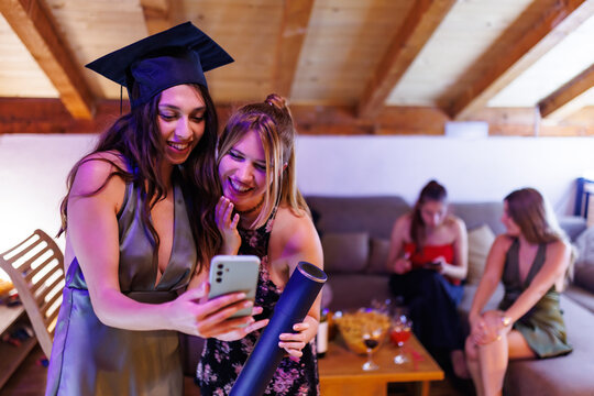 Two friends celebrating a graduation party and capturing joyful moments on a smartphone in a warm and cozy setting