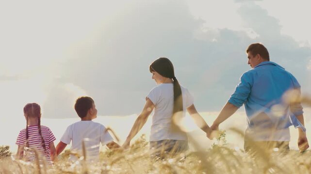 Happy family holding hands walking at bright sunny dry wheat field back view. Mother father son and daughter with love going cereal harvest plantation natural sunlight sky horizon agriculture scenery