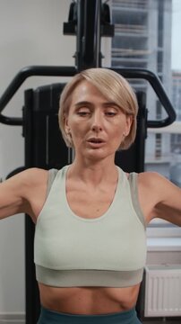 Vertical shot of adult Caucasian female athlete performing chest fly movement while sitting in pec deck machine in modern gym