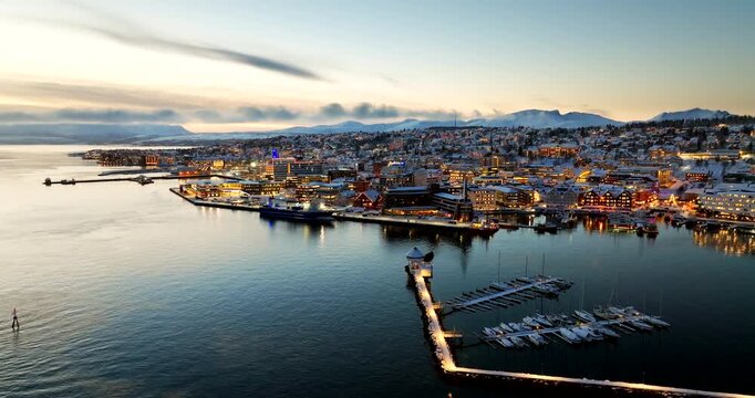 Aerial establishing shot of Tromso city in Norway, covered in snow during blue hour. Features illuminated harbor, marina, and snowy mountains