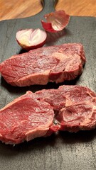 Obraz premium Raw Steaks With Garlic Clove Accents On Dark Board, Aromatic Infusion Prep By Backyard Chef, Rustic Lighting Highlighting Moist Surface And Savory Promise, Ready For Searing