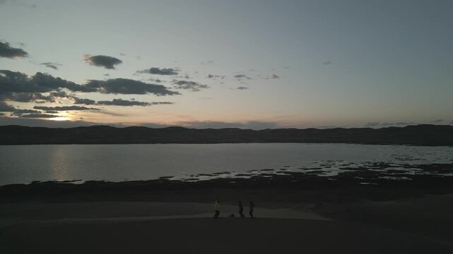 Raw drone footage of group of friends jumping and waving hands on top of sand dune during sunset in Mongolia, aerial view of travelers at golden hour with lake and desert background, unedited.