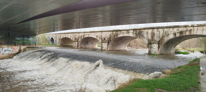 Puente antiguo de arcos de piedra bajo una estructura moderna con una ca&iacute;da de agua espumosa en el r&iacute;o en Burgos, Espa&ntilde;a