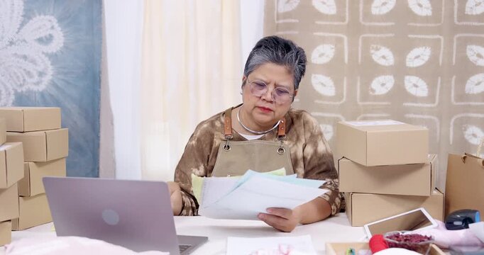 Senior asian woman entrepreneur reads invoice packet, checks online order list on laptop, compares paperwork, plans parcel packing steps, prepares shipping decisions inside home studio. home studio