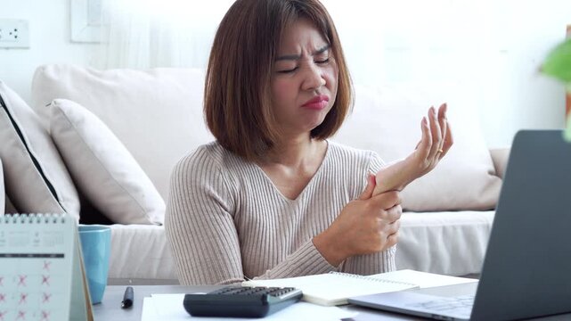Asian woman suffering from finger joint inflammation and hand pain while working on a laptop at home. Female freelancer feeling office syndrome symptoms or arthritis after long hours of typing
