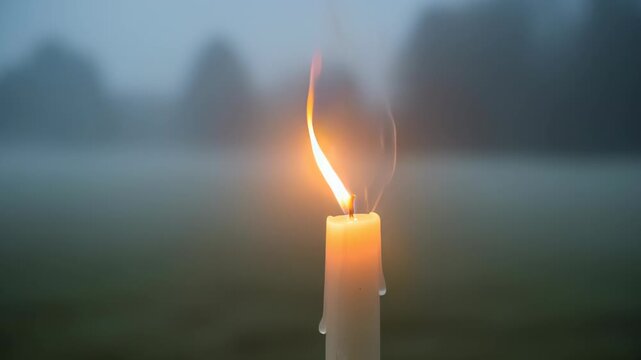 Close-up of a single lit candle with flickering flame in a foggy outdoor setting during early morning or evening, symbolizing warmth and spiritual reflection for Imbolc