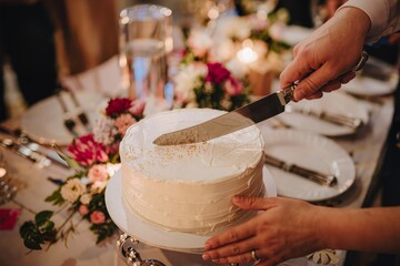 Close up of a couple cutting a simple white frosted wedding cake with a silver knife during a romantic reception dinner with colorful flowers and warm candlelight in the blurred background.