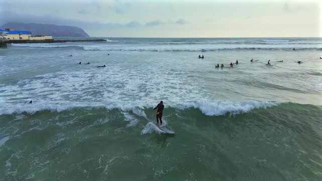 Long board surfer surfing large left wave break Miraflores Playa Makaha Beach aerial drone Lima Peru Barranquito sunset golden hour haze clouds crowds surf school white wash backwards pan follow
