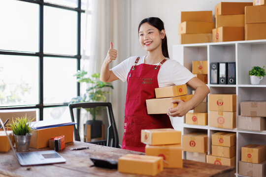 Asian woman sme business owner wearing a brown apron holding boxs is smiling and giving thumbs up while standing in a warehouse full of cardboard boxes
