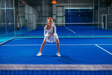 Obraz premium Female padel player in white outfit ready to return a shot on a blue indoor court