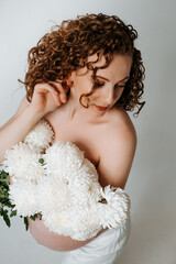 Fototapeta premium a pregnant woman with flowers on a white isolated background patting her belly and enjoying her pregnancy, a young girl with a big belly at home waiting for a baby, showing a heart with her hands
