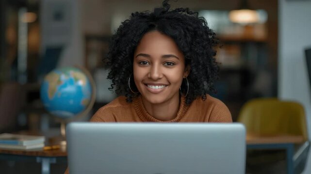 Smiling young woman with curly hair at cozy study workspace working on laptop and study materials