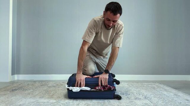 Young man having difficulty closing his luggage, forcing the lid shut on the overpacked suitcase as he prepares for a trip or vacation