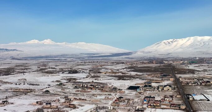 A snowy Aragats mountain range is in the background of a town. The town is small and has a few houses. The sky is clear and blue