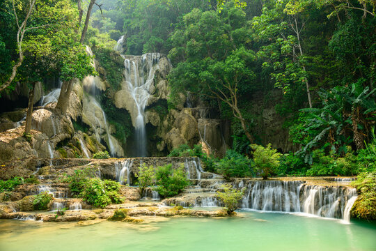 Limestone cascades flow over multiple levels surrounded by dense tropical foliage at Kuang Si Waterfalls in northern Laos. Sunlight highlights the turquoise pools and lush greenery, creating a vibrant