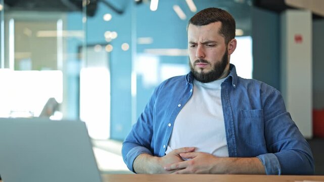 Individual experiences discomfort and stomach pain while concentrating on laptop work in modern office setting. Unwellness and indigestion symptoms cause distress, interrupting productivity.