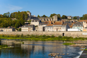 Obraz premium Ancient stone ramparts and round towers rise above the traditional tiled rooftops of La Charite sur Loire, with the Loire river and lush greenery in the foreground. Warm afternoon sunlight highlights
