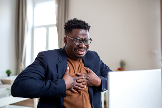 African American businessman experiencing sudden chest pain or heart attack while working at a laptop. Man looking distressed and holding his chest, suffering from pressure, anxiety, or heartburn.