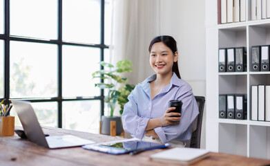 Young asian woman working remotely at office, holding a disposable coffee cup, sipping coffee while working on her laptop at a table. people and lifestyle concepts
