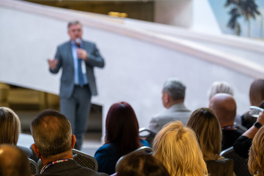 Speaker in suit delivers talk to attentive crowd outdoors near modern venue daylight