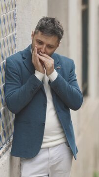 A man in a blue jacket stands against the wall and plays the harmonica in the street