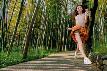 Obraz premium Woman jump along a bamboo path in a forest, casual summer outfit and smile, motion captured midair on a sunny outdoor walkway conveying joyful energy and carefree lifestyle.