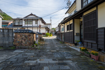 Obraz premium Charming narrow alley lined with stone and plaster walls, wooden facades, and potted flowers in Arita, Japan. Morning light highlights the quiet residential street beneath a clear sky, with green