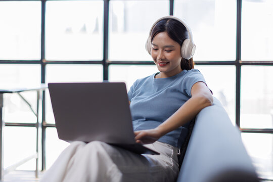 Asian cute girl wearing headphones and using laptop enjoying free time sitting on a sofa at home
