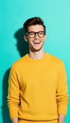 Fototapeta premium A joyful young man wearing a bright yellow sweater and stylish glasses laughs heartily, looking directly at the camera against a vibrant turquoise background.