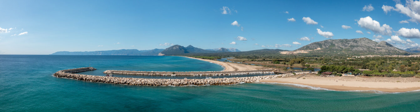 Panoramic aerial view of a curved stone breakwater enclosing turquoise waters, sandy beach, and green countryside with rugged mountains under a clear blue sky near Orosei, Sardinia.