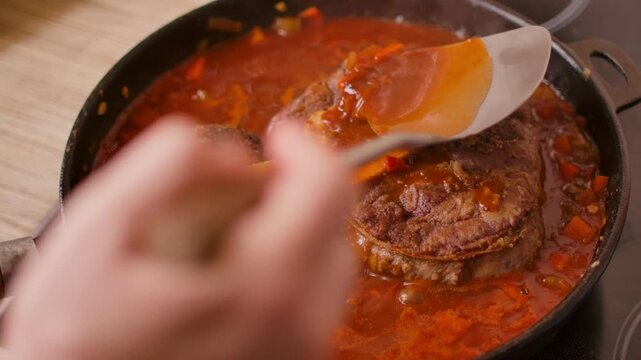 A chef uses a ladle to pour hot vegetable sauce over osso buco. veal shanks simmering in a pan The rich gravy coats the meat during the slow cooking process for a gourmet meal.