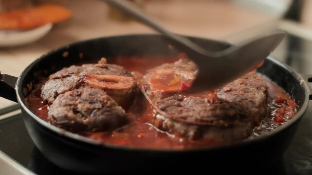 A chef uses a ladle to pour hot vegetable sauce over osso buco. veal shanks simmering in a pan The rich gravy coats the meat during the slow cooking process for a gourmet meal.