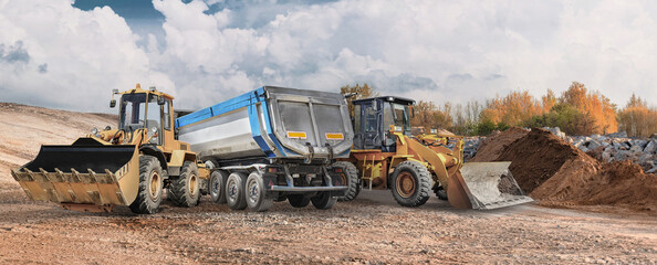 Loader and excavator are at a construction site moving dirt. A dump truck waits to be loaded with material. The work is taking place in a wooded area © Anoo