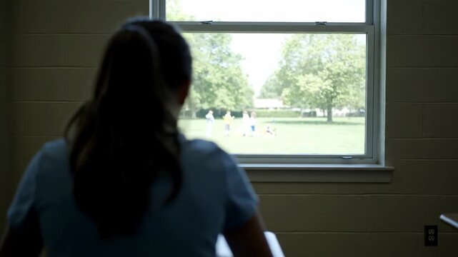 Rear view of a female student looking out the classroom window. Lonely girl watching children play outside during school