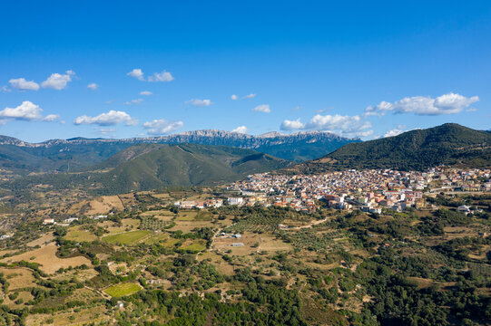 Aerial photo of Orgosolo perched on rolling hills, surrounded by patchwork fields and dramatic mountain peaks under a clear blue sky in Sardinia.