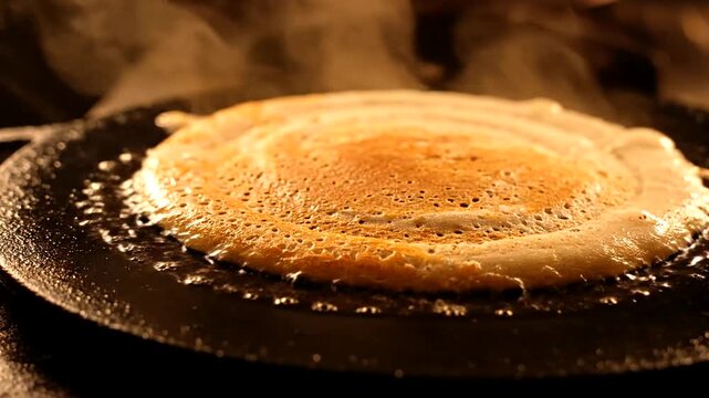 Close-up of a Dosa cooking on a hot griddle, steam rising.