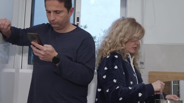 Mature couple having a distant morning routine, man engrossed in his phone while his wife cooks breakfast in the kitchen, feeling neglected and showing indifference