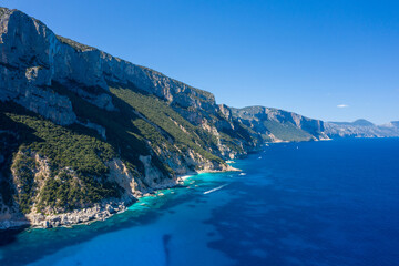 Obraz premium Towering limestone cliffs covered with dense green vegetation rise sharply above the deep azure waters of the Mediterranean near Cala Goloritze, Baunei, Sardinia. Bright sunlight and clear skies
