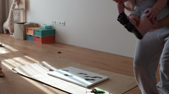 Selfie UGC video of parents holding a baby and showing a paper with footprint painting. Black baby footprints visible on the sheet during a diy activity at home.

