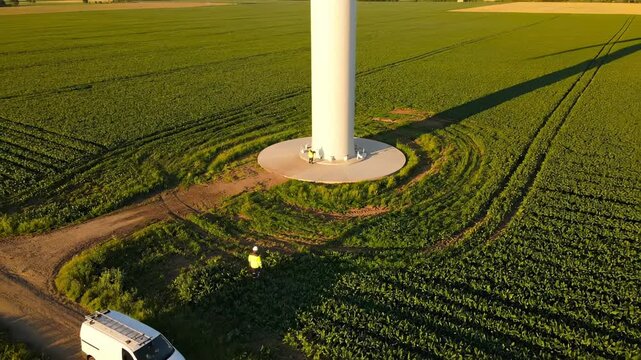 Wind turbine stands in green field with technician inspecting base. Worker maintains turbine in agricultural field. Wind energy technician works near turbine. Service van parked at wind tower.
