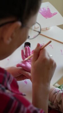 mother painting a toddler&rsquo;s hand with a brush during a simple art activity at a table. close up view of adult hands and child&rsquo;s hand. indoor home setting