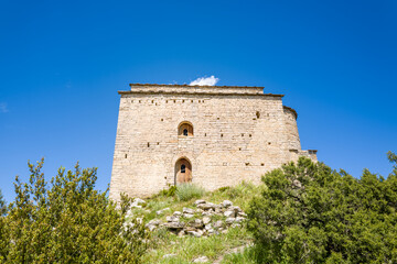 Historic stone castle stands atop a grassy hill in Samitier, Spain, its weathered facade illuminated by bright sunlight against a vibrant blue sky and surrounded by green shrubs. © Florent