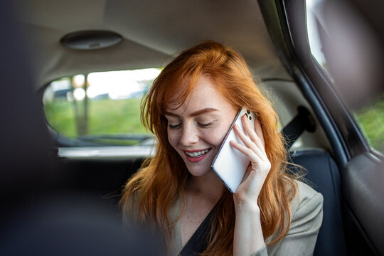 Happy young redhead woman talking on smartphone in back seat of car. Ginger girl passenger using mobile phone and smiling while traveling in taxi. Communication and technology lifestyle concept.