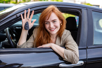 Happy young redhead woman showing car key through vehicle window. Smiling female driver holding remote key for new automobile. Buying a car, rental service, dealership and auto insurance concept info.
