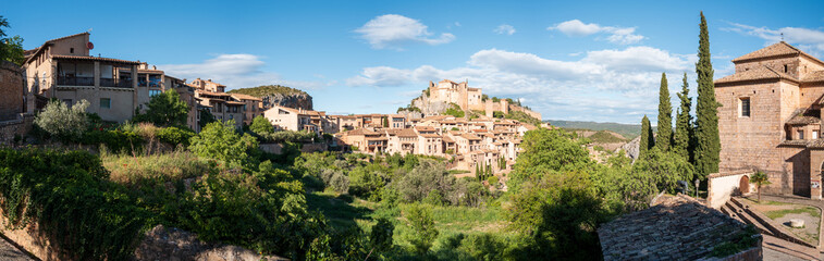 Obraz premium Wide-angle scene of Alquezar, Spain, showing historic stone houses, cypress trees, and the prominent castle atop a hill under a bright blue sky. Lush greenery and textured rooftops add depth to the