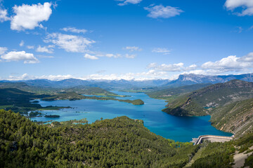 Fototapeta premium Expansive aerial landscape of Mediano Lake in Aragon, Spain, featuring turquoise water, lush green hills, and distant snow-capped Pyrenees mountains beneath a vivid blue sky with scattered clouds.