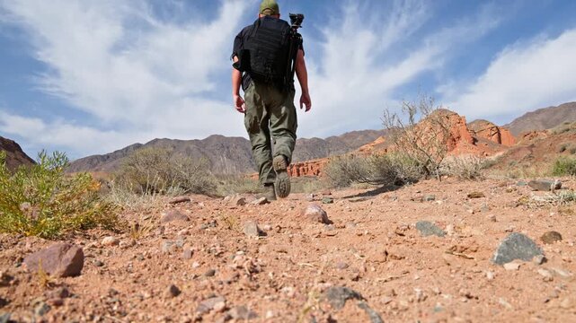 Determined man explores desert landscape while hiking. Adult Caucasian with backpack sets up camera on tripod, embodying adventure spirit.
