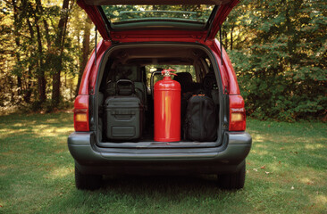 Red fire extinguisher with a smooth, glossy surface and cylindrical shape placed in the open trunk of a red suv, accompanied by black travel bags, set in a lush green outdoor