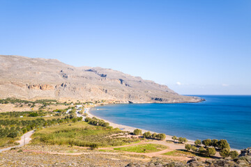 A bright panoramic view captures the curved sandy bay and turquoise waters of Kato Zakros, Crete, framed by arid rocky hills and scattered greenery under a clear blue sky. The scene radiates calm and