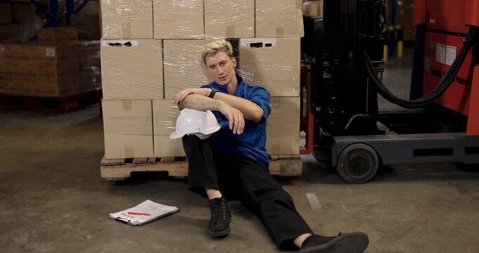 caucasian male logistic warehouse worker sitting on floor leaning against stacked boxes holding helmet during break looking ahead with calm exhausted expression after working in storage facility